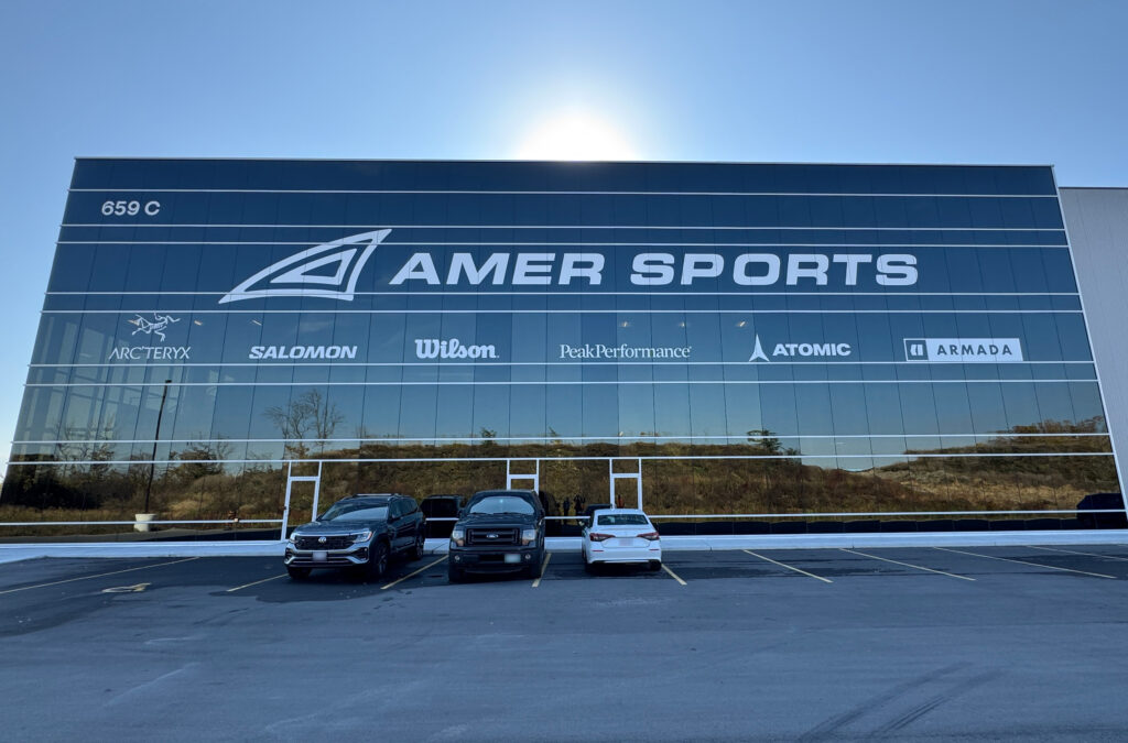 Exterior view of Amer Sports' building featuring brand logos, with cars parked in front under a clear blue sky.